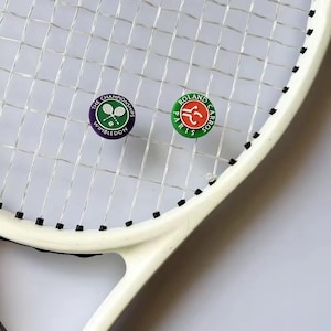 Op de afbeelding: Een witte tennisracket met een snarennet, met twee ronde pins. Eén pin toont het Wimbledon-logo in groen en paars, de andere het Roland Garros-logo in rood en groen. De racket staat tegen een lichte achtergrond.