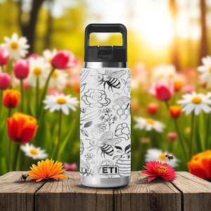 Puede incluir: Una botella de agua de acero inoxidable con asa y tapa negras. El cuerpo blanco presenta un patrón de abejas y flores en negro y gris. La botella está sobre una superficie de madera con flores en el fondo.