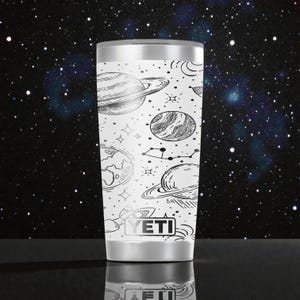May include: A white YETI tumbler featuring a black line art pattern of various planets, stars, and constellations. The cylindrical drinkware has a metallic rim and base, with the YETI brand name clearly printed in a rectangular box at the bottom.