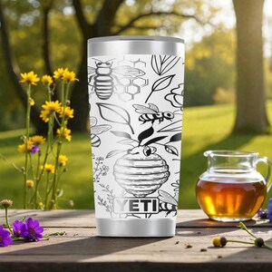 May include: A white stainless steel tumbler featuring a black line art pattern of bees, honeycombs, leaves, and a beehive. The YETI brand logo is printed in black at the bottom center of the cylindrical container, which has a metallic silver rim.