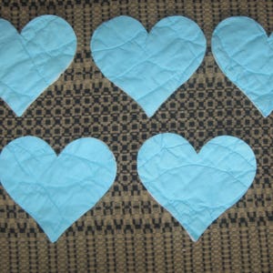 May include: Five light blue quilted fabric hearts arranged in two rows. A white ruler is positioned on the left side, showing each heart measures approximately 4 inches in height and width. The hearts feature a stitched grid pattern.