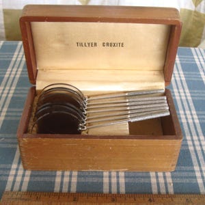 May include: A set of five circular dark-tinted glass lenses with silver-toned metal handles, arranged in a wooden storage box. The interior lid features the text TILLYER CRUXITE. A wooden ruler is visible at the bottom, showing a 5-inch scale.