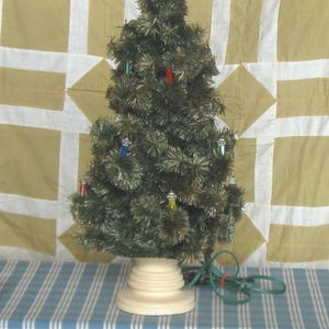 May include: A tabletop artificial Christmas tree with dark green tinsel needles, featuring small multi-colored light bulbs and a silver star topper. The tree is supported by a round, off-white pedestal base and includes a green electrical cord.