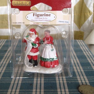 May include: A Christmas figurine set in a clear plastic package. The set includes a Santa Claus figure in a red suit and a woman in a red dress and white apron. The package has the text "Figurine Chocolate To Go".