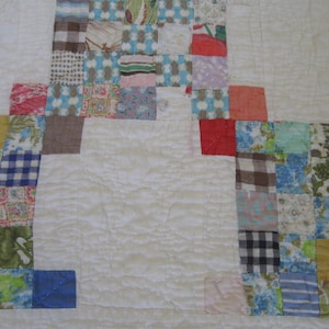 May include: A patchwork quilt featuring a central large cream-colored square surrounded by a border of smaller, multi-colored fabric squares. The quilt displays various patterns including gingham, floral, and striped prints in blue, red, green, and brown.