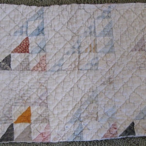 May include: A rectangular quilted fabric piece featuring a repeating pattern of triangular blocks in various muted colors including blue, grey, brown, and orange, set against a white background with a diamond-shaped stitched texture throughout.
