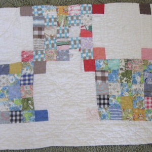 May include: A patchwork quilt with a white background and colorful squares. The squares are in various patterns and colors, including blue floral, red gingham, and brown plaid. The quilt has a simple, classic design.