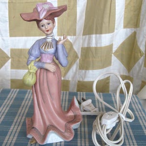May include: A porcelain figurine lamp shaped like a person wearing a wide-brimmed pink hat, a lavender long-sleeved top with a gold-accented collar, and a long pink skirt. The figure holds a yellow pouch and includes a white electrical cord.