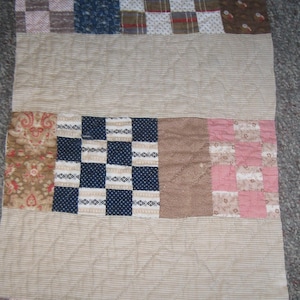 May include: A rectangular patchwork quilt featuring alternating horizontal bands of solid beige striped fabric and sections of various patterned squares. The patterns include navy blue polka dots, pink and white checks, floral prints, and plaid designs.