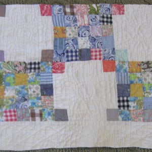 May include: A rectangular quilt featuring a white background with three distinct blocks of multi-colored fabric squares. The squares display various patterns including floral, plaid, and gingham prints in shades of blue, yellow, pink, and green.