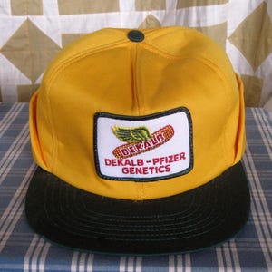 May include: A yellow baseball cap with a dark green brim. The cap features a patch with the text "DEKALB - PFIZER GENETICS" and a winged corn graphic. The cap has a green button on top.