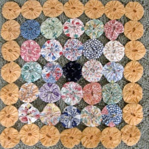 May include: A square quilt featuring a repeating pattern of fabric circles. The circles are in various floral patterns and colors, including blue, red, and pink. The border circles are a solid light orange color.