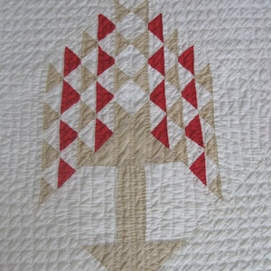 May include: A square quilted wall hanging featuring a central tree motif constructed from geometric fabric pieces in red, beige, and white. The design uses triangular and diamond shapes to form the tree canopy, with a solid beige trunk and base.