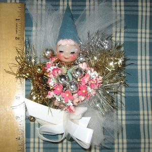 May include: A decorative elf ornament with a blue felt hat, pink cheeks, and closed eyes. It is adorned with gold and silver tinsel, pink flowers, and silver spheres, finished with a white tulle bow. A wooden ruler shows it is 6 inches tall.
