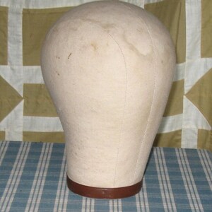 May include: A beige canvas-covered mannequin head with visible seams along the crown and sides. The head is mounted on a circular, dark brown base. The surface of the fabric shows light signs of wear and discoloration across the top and sides.
