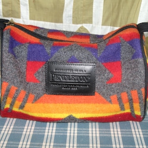 May include: A rectangular wool pouch featuring a geometric pattern in shades of orange, red, purple, and grey. The bag has a black zipper closure and a black leather patch centered on the front with the text Warranted to be a Pendleton.