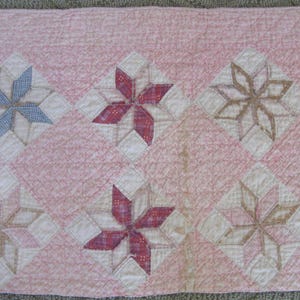May include: A vintage quilt with a pink background and six starburst patterns. The starbursts are in shades of blue, red, and brown, set within white diamond shapes. The quilt is hand-stitched.