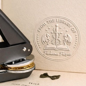 May include: A dark gray seal press with a gold and silver seal, next to a cream-colored book with a raised seal that reads "FROM THE LIBRARY OF Farheena Faiyaz." A small, decorative metal accent is also present.