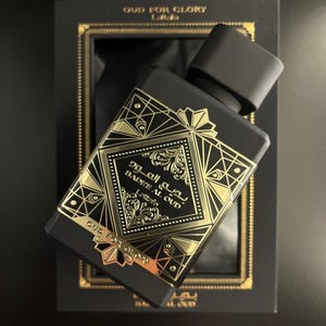 May include: Black perfume bottle with gold geometric designs and the text "BADEE AL OUD" and "OUD FOR GLORY". The bottle is angled in a black box with gold trim. The bottle's cap is black.