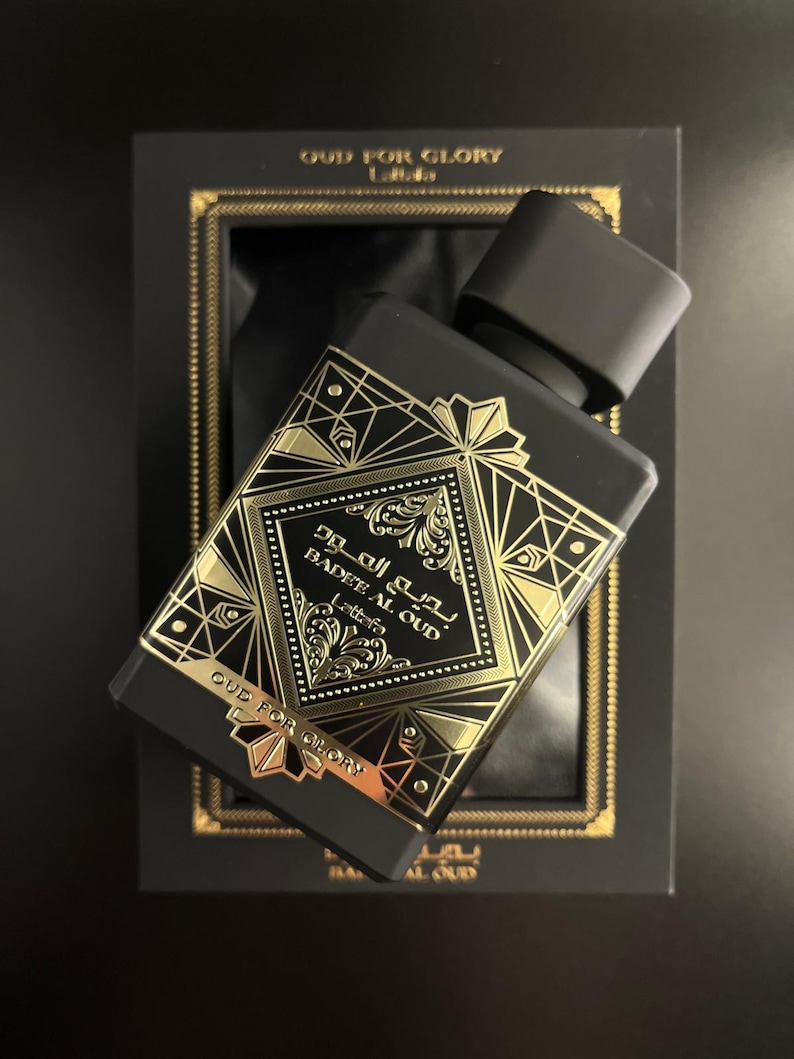 May include: Black perfume bottle with gold geometric designs and the text "BADEE AL OUD" and "OUD FOR GLORY". The bottle is angled in a black box with gold trim. The bottle's cap is black.