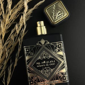 May include: Black rectangular perfume bottle with gold accents and a gold cap. The bottle has gold geometric designs and text that reads "Badee Al Oud" and "Oud for Glory". The bottle is next to a gold-topped square cap.