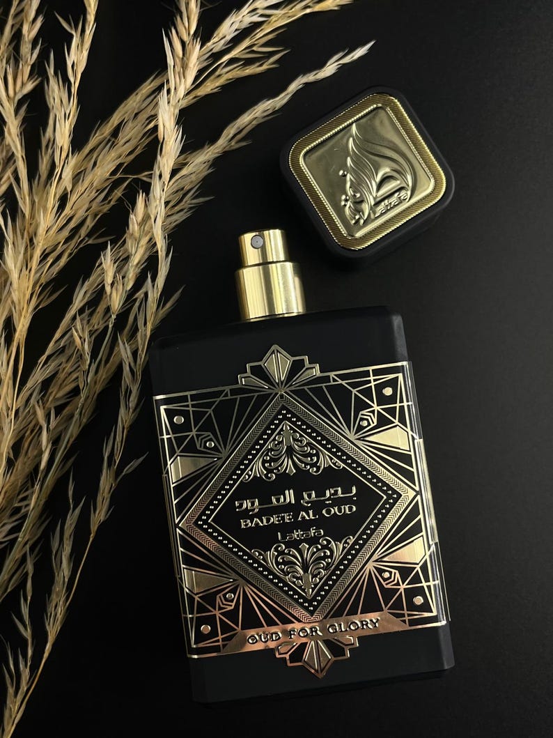 May include: Black rectangular perfume bottle with gold accents and a gold cap. The bottle has gold geometric designs and text that reads "Badee Al Oud" and "Oud for Glory". The bottle is next to a gold-topped square cap.