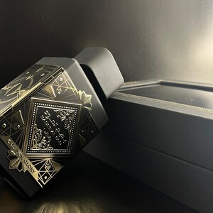 May include: Black perfume bottle with gold geometric designs and Arabic script. The bottle is next to a black box with gold trim. The text "OUD FOR GLORY" is visible on the bottle.