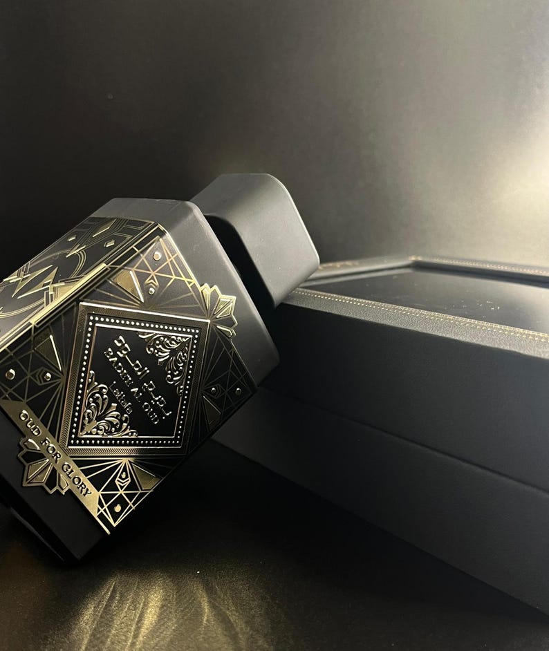 May include: Black perfume bottle with gold geometric designs and Arabic script. The bottle is next to a black box with gold trim. The text "OUD FOR GLORY" is visible on the bottle.