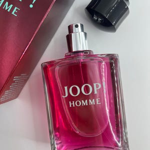Puede incluir: Un frasco de perfume rectangular de vidrio lleno de un líquido rosa vibrante. El frasco tiene un atomizador plateado y las palabras "JOOP! HOMME" están impresas en blanco. Una tapa negra está al lado del frasco y una caja burdeos es parcialmente visible.