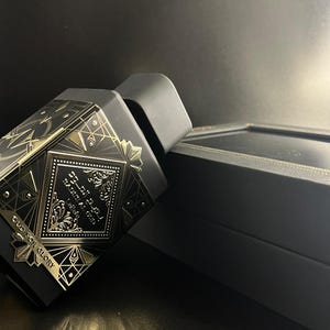 May include: Black perfume bottle with gold geometric designs and Arabic script. The bottle is next to a black box with gold trim. The text "OUD FOR GLORY" is visible on the bottle.