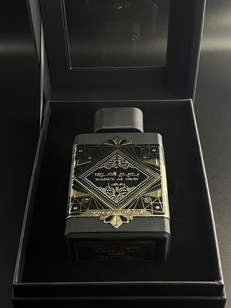 May include: A rectangular black perfume bottle with gold accents and the text "BADEE AL OUD" and "OUD FOR GLORY". The bottle is in a black box with a clear lid. The design features geometric patterns and Arabic script.