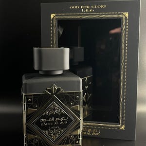 May include: A black perfume bottle with gold accents and the text "BADEE AL OUD" and "OUD FOR GLORY". The bottle is rectangular with a black cap and is displayed in a black box with gold trim.