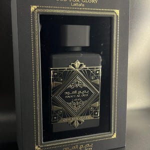 May include: A black perfume bottle with gold accents is displayed inside a black box. The bottle has a geometric design with Arabic script and the words "OUD FOR GLORY" and "BADEE AL OUD" in gold. The box also features gold trim.