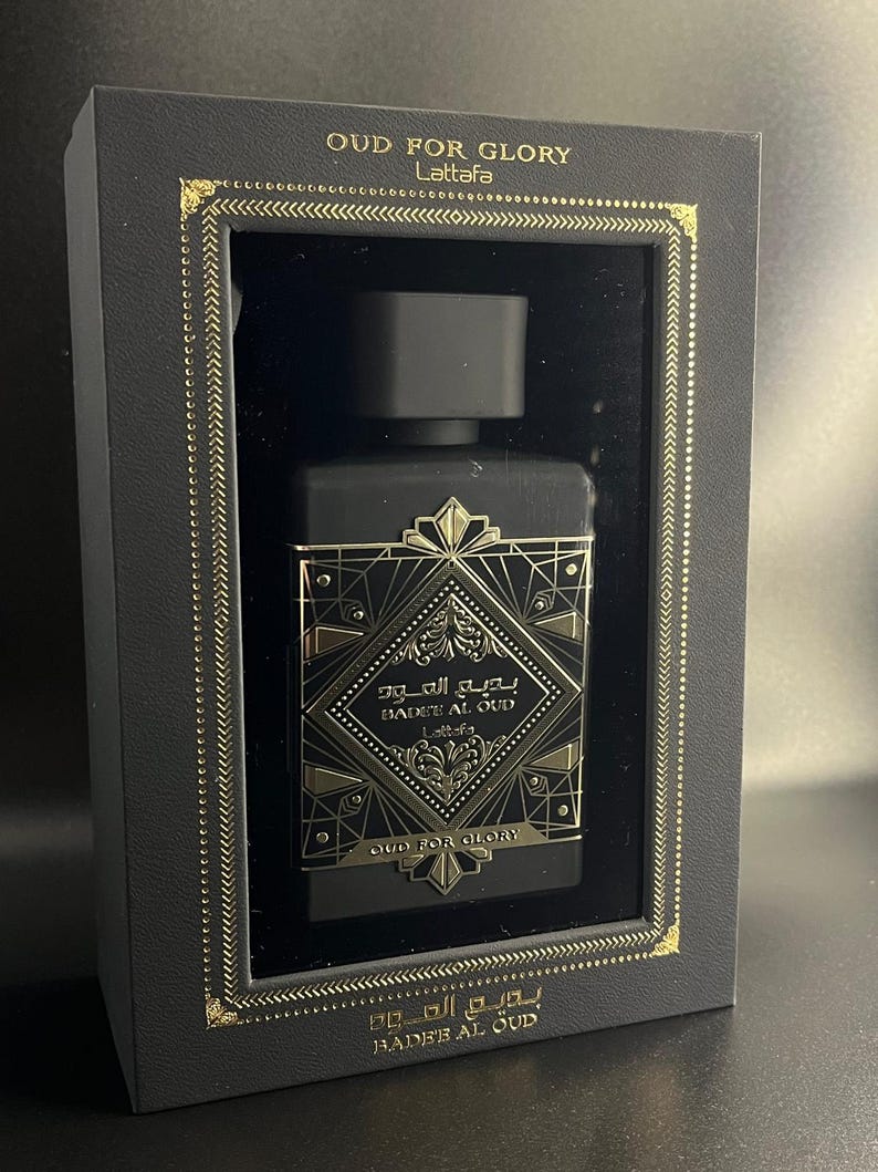 May include: A black perfume bottle with gold accents is displayed inside a black box. The bottle has a geometric design with Arabic script and the words "OUD FOR GLORY" and "BADEE AL OUD" in gold. The box also features gold trim.