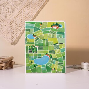 May include: A rectangular notebook featuring a vibrant, illustrated cover design of a patchwork landscape. The artwork displays various shades of green fields, small clusters of trees, several blue ponds, and tiny red-roofed buildings.