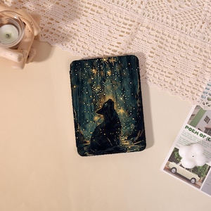 May include: A rectangular e-reader case featuring a dark forest scene with a black cat sitting and looking up at glowing golden stars and light particles. The design is set against a deep green and black background with vertical tree trunks.