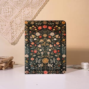 May include: A rectangular journal with a dark green floral pattern. The design features red, yellow, and white flowers with green leaves. The journal is standing upright on a white surface.