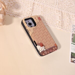May include: A phone case with a brown floral pattern and a silver frame. The case features a cartoon character with a red bow and autumn leaves. The design is set against a light beige background.