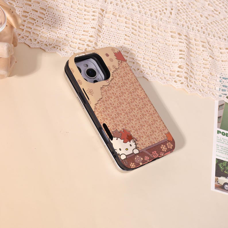 May include: A phone case with a brown floral pattern and a silver frame. The case features a cartoon character with a red bow and autumn leaves. The design is set against a light beige background.