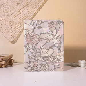 Könnte beinhalten: Eine rechteckige Tablethülle mit einem Design im Buntglasstil, das zartrosa und weiße Rosen, geschwungene Stängel und Blätter zeigt. Die Oberfläche hat ein irisierendes, perlmuttartiges Finish mit dunklen Umrandungen.