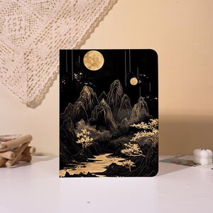 Puede incluir: Un cuaderno rectangular con una cubierta negra que presenta una ilustración en tonos dorados de picos montañosos escarpados, un río sinuoso y árboles bajo un cielo nocturno con dos lunas llenas y rayos de luz verticales descendentes.
