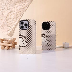 Op de afbeelding: Twee telefoonhoesjes met een Snoopy-ontwerp. De hoesjes zijn gebroken wit met zwarte stippen en een cartoon Snoopy-illustratie. De ene hoes is groter dan de andere, met een uitsparing voor een drievoudige cameralens. De hoesjes zijn gemaakt van een glad materiaal.