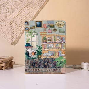 May include: A decorative panel featuring a cozy interior scene with Snoopy reading a book in a green armchair. A bookshelf, a clock, and the text "you're doing a really good job" are also visible. The color palette includes greens, blues, and browns.