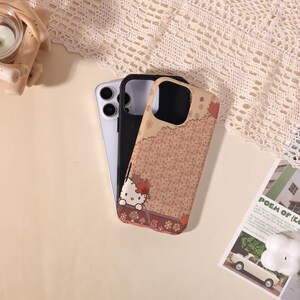 May include: Three phone cases: a silver, a black, and a tan case with a Hello Kitty design. The tan case features a floral pattern and a Hello Kitty graphic with fall leaves. The cases are on a cream-colored surface.