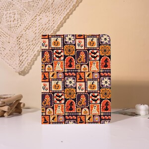 May include: A rectangular notebook featuring a repeating pattern of Halloween-themed icons including orange pumpkins, black cats, white ghosts, bats, spider webs, mushrooms, and witch hats set against a dark purple and black background.