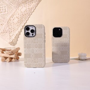 May include: Two beige phone cases with a repeating geometric tile pattern. The cases have black camera cutouts and side buttons. The cases are displayed on a white surface with a neutral background.