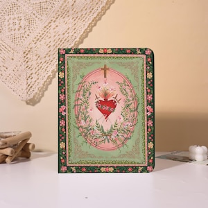 Peut inclure: Un journal décoratif avec une bordure verte et un motif central représentant un cœur rouge percé de trois épées, entouré de fleurs et de feuillage. Une croix dorée est en haut, sur un ovale rose. Le journal est sur une surface blanche.