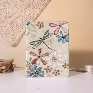 May include: A decorative tablet case featuring a dragonfly and floral design. The case has a light green background with colorful flowers in shades of pink, blue, and red. The dragonfly is black and green.