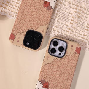 May include: Two phone cases with a tan and brown floral pattern and a torn paper design. One case features a white cat face with red and brown leaves. The cases have black camera cutouts.