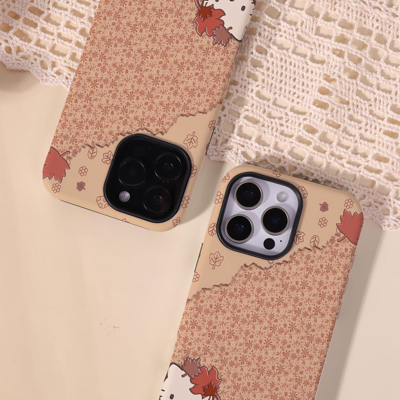 May include: Two phone cases with a tan and brown floral pattern and a torn paper design. One case features a white cat face with red and brown leaves. The cases have black camera cutouts.